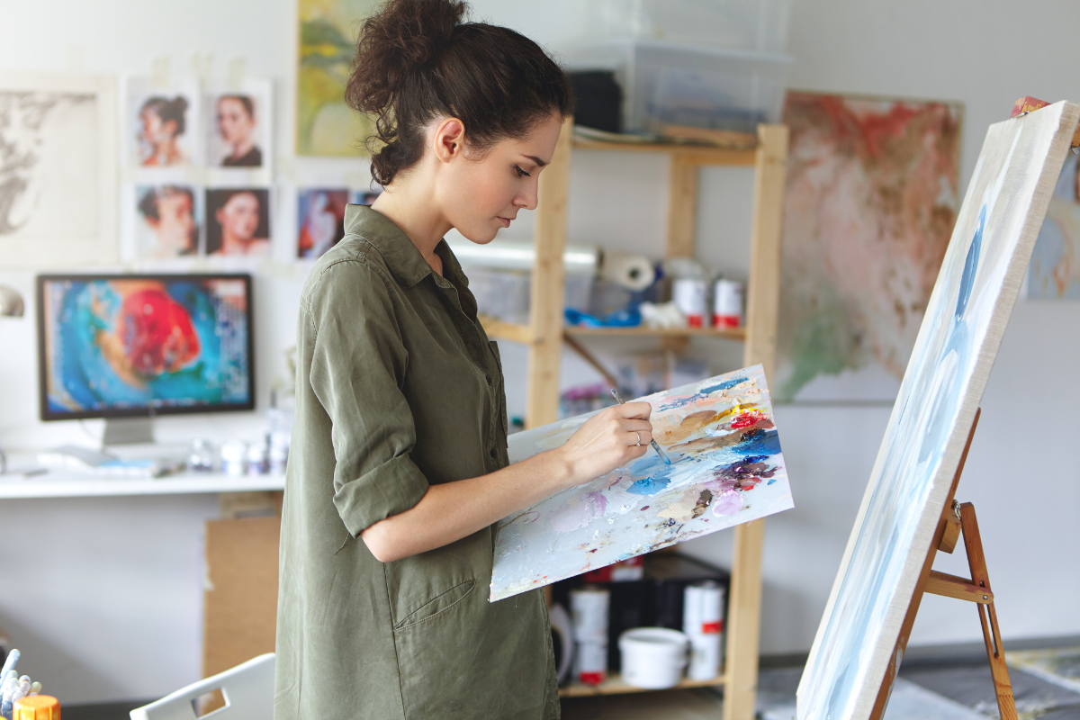 estudante de artes visuais ead pintando uma tela branca. ela segura uma placa com tintas, um pincel, está com cabelo preso e veste uma camisa verde militar.